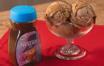 Fotografia de uma taça de vidro com três bolas de sorvete de café. Ao lado tem uma embalagem de NESCAFÉ Gelado, sobre um pano vermelho, apoiado em uma mesa de madeira.