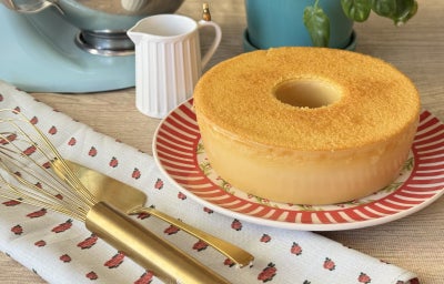 Foto da receita de bolo mole cearense, servido em um prato colorido, sobre uma mesa de café da tarde, com talheres dourados, uma batedeira azul ao fundo