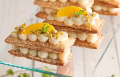 Fotografia em tons de amarelo em uma bancada de madeira com um suporte para bolo de vidro e quatro mil folhas com recheio de creme e pêssegos.