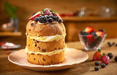 Fotografia em tons de marrom de uma bancada marrom com um prato rosa, sobre ele um panettone recheado com frutas vermelhas no topo. Ao lado um recipiente de vidro com frutas vermelhas. Fotografia em tons de marrom de uma bancada marrom com um prato rosa, sobre ele um panettone recheado com frutas vermelhas no topo. Ao lado um recipiente de vidro com frutas vermelhas.