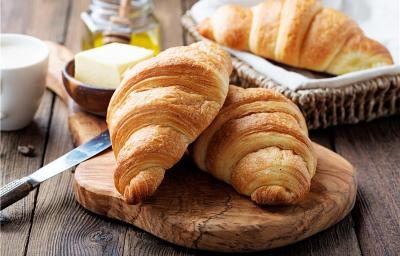 Fotografia de dois croissants sobre uma pequena tábua de madeira, sobre uma mesa também de madeira, mas em tom escuro. Apoiado na tábua tem uma faca e ao fundo, tem outro croissant dentro de uma cestinha com um pano branco. Fotografia de dois croissants sobre uma pequena tábua de madeira, sobre uma mesa também de madeira, mas em tom escuro. Apoiado na tábua tem uma faca e ao fundo, tem outro croissant dentro de uma cestinha com um pano branco.