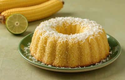 Fotografia de um bolo bundt de milho, dourado e coberto com coco, apresentado em um prato verde, com espigas de milho e uma lima ao fundo.