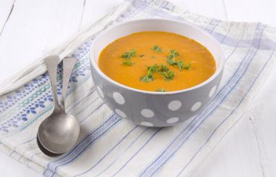 fotografia em tons de branco, laranja e azul de uma bancada branca vista de cima. Contém um pano branco com listras azuis, por cima um recipiente redondo cinza com bolinhas brancas e dentro a sopa. Ao lado do recipiente duas colheres. fotografia em tons de branco, laranja e azul de uma bancada branca vista de cima. Contém um pano branco com listras azuis, por cima um recipiente redondo cinza com bolinhas brancas e dentro a sopa. Ao lado do recipiente duas colheres.