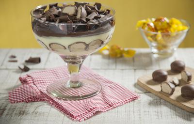 Fotografia em tons de amarelo e chocolate, ao centro taça com creme, bombons cortados ao meio e raspas de chocolate sobre guardanapo xadrez branco e vermelho, ao lado tábua com bombons, ao fundo potinho com bombons, tudo em bancada de madeira em pátina.