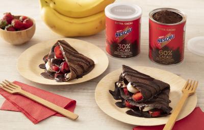 Foto em tons de vermelho da receita de crepioca doce feita com Nescau extra cacau servida em duas porções em pratos de madeira sobre panos vermelhos. Ao fundo, morangos e bananas de decoração, além das latas de nescau e garfos de madeira Foto em tons de vermelho da receita de crepioca doce feita com Nescau extra cacau servida em duas porções em pratos de madeira sobre panos vermelhos. Ao fundo, morangos e bananas de decoração, além das latas de nescau e garfos de madeira