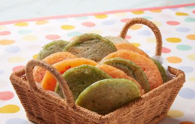 Fotografia em tons de verde e laranja em uma bancada de madeira, uma toalha branca com bolinhas coloridas, uma cesta de vime marrom clara com pães de queijo achatados nas cores verde e amarelo. Fotografia em tons de verde e laranja em uma bancada de madeira, uma toalha branca com bolinhas coloridas, uma cesta de vime marrom clara com pães de queijo achatados nas cores verde e amarelo.