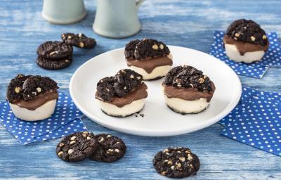 Foto de uma bancada de madeira azulada, com guardanapos azuis de bolinhas brancas espalhados e, ao centro, um prato branco com três mini cheesecakes decoradas com Cookies Negresco. Há mais cookies Negresco espalhados pela bancada Foto de uma bancada de madeira azulada, com guardanapos azuis de bolinhas brancas espalhados e, ao centro, um prato branco com três mini cheesecakes decoradas com Cookies Negresco. Há mais cookies Negresco espalhados pela bancada