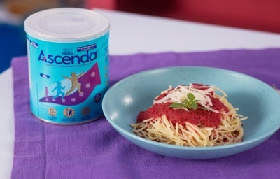 Fotografia de um prato fundo azul, pequeno, sobre um pano roxo, ao lado de uma lata de Ascenda. Dentro do prato tem macarrão tradicional com o molho de tomate e ascenda.