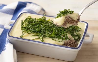Fotografia em tons de azul em uma bancada e madeira, um pano xadrez azul e ao centro um recipiente refratário branco com detalhes em azul e o escondidinho de carne e batata e couve por cima para decorar.