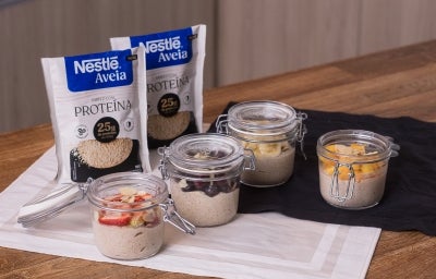 Receita de overnight oats proteico para a semana servida em potes herméticos de vidro sobre uma mesa de madeira decorada com panos e embalagens de aveia proteína