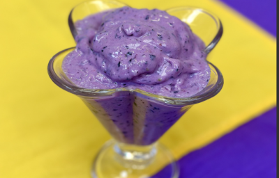 sobre um fundo azul, amarelo e branco, dentro uma taça de vidro transparente está um sorvete cremoso em tons de roxo com pedacinhos de uva.