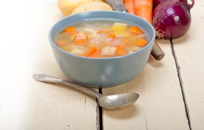 Fotografia de uma sopa de batata, cenoura e chuchu, dentro de um recipiente de vidro na cor azul. Na frente, uma colher de sopa, e ao fundo, cebolas roxa, batatas e cenoura, sobre uma mesa de madeira. Fotografia de uma sopa de batata, cenoura e chuchu, dentro de um recipiente de vidro na cor azul. Na frente, uma colher de sopa, e ao fundo, cebolas roxa, batatas e cenoura, sobre uma mesa de madeira.