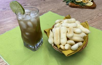 Fotografia em tons de verde em uma bancada de madeira, um pano verde, uma cesta de vinil com guardanapo amarelo com os biscoitos de polvilho em cima, ao lado um copo de vidro alto com o chá verde, cubos de gelo e uma rodela de limão decorando. Fotografia em tons de verde em uma bancada de madeira, um pano verde, uma cesta de vinil com guardanapo amarelo com os biscoitos de polvilho em cima, ao lado um copo de vidro alto com o chá verde, cubos de gelo e uma rodela de limão decorando.