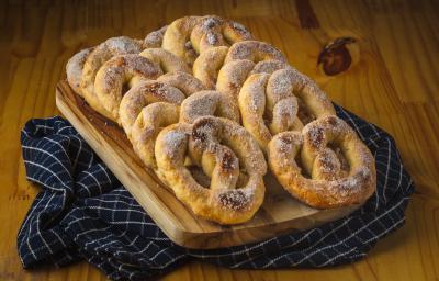 Foto aproximada de uma bancada de madeira sobre a qual está um pano azul quadriculado e nele há uma tábua de madeira com alguns pretzels decorados com açúcar e canela