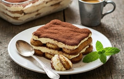 Em cima de uma mesa, prato com porção de tiramisu feito com biscoitos champanhe, creme branco cremoso, polvilhado com cacau em pó.