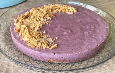 Fotografia mostra uma cheesecake de cor roxa devido ao açaí, com uma farofa de biscoito maisena no topo.