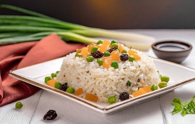 Em um prato branco, porção de arroz branco com uvas-passas, ervilha e cenouras picadas em cubos.