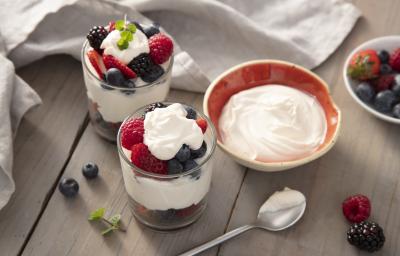 Fotografia em tons de vermelho em uma bancada de madeira clara, um pano cinza, um potinho com o mascarpone, um potinho com frutas vermelhas. Ao centro, dois copos de vidro com frutas vermelhas cobertas por marcarpone.