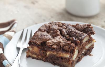 Fotografia em tons de azul e marrom em uma bancada de madeira clara, um pano listrado em azul e marrom, um prato branco redondo raso com um pedaço de torta de banana com chocolate.