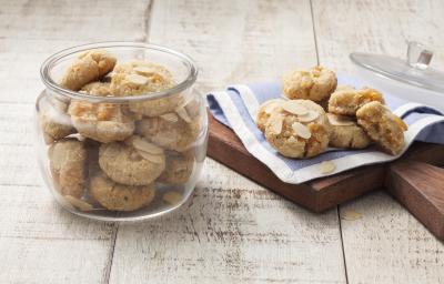 Fotografia em tons de azul em uma bancada de madeira clara com um pano azul sobre uma tábua de madeira com alguns cookies de amêndoas e damasco apoiados nele. Ao lado, um pote grande de vidro com mais cookies dentro. Fotografia em tons de azul em uma bancada de madeira clara com um pano azul sobre uma tábua de madeira com alguns cookies de amêndoas e damasco apoiados nele. Ao lado, um pote grande de vidro com mais cookies dentro.
