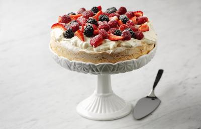 Fotografia em tom claro, com uma torta recheada com suspiro e frutas por cima, polvilhadas com açúcar de confeiteiro. A torta está apoiada sobre um prato alto branco decorativo, ao lado, uma espátula prata para bolo, tudo sobre uma mesa de mármore branca. Fotografia em tom claro, com uma torta recheada com suspiro e frutas por cima, polvilhadas com açúcar de confeiteiro. A torta está apoiada sobre um prato alto branco decorativo, ao lado, uma espátula prata para bolo, tudo sobre uma mesa de mármore branca.