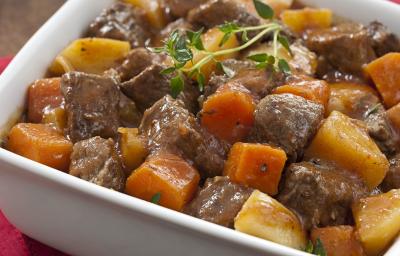Fotografia em tons de marrom com um paninho vermelho e um recipiente branco retangular com picadinho de carne com batatas e cenoura