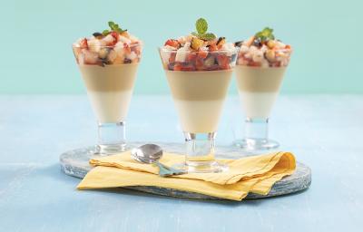 Foto em tons de azul da receita de pudim de coco com calda de ameixa servida em 3 taças de vidros com pedaços das frutas por cima, as taças sobre uma tábua de vidro com um pano amarelo e uma colher prateada para decoração. Foto em tons de azul da receita de pudim de coco com calda de ameixa servida em 3 taças de vidros com pedaços das frutas por cima, as taças sobre uma tábua de vidro com um pano amarelo e uma colher prateada para decoração.