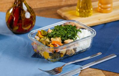 Fotografia de uma marmita retangular pequena com arroz, feijão, carne suína, couve e legumes assados. A marmita está sobre dois panos sobrepostos de cor azul, ao lado de talheres e uma mini tábua com pimenta e azeite. Fotografia de uma marmita retangular pequena com arroz, feijão, carne suína, couve e legumes assados. A marmita está sobre dois panos sobrepostos de cor azul, ao lado de talheres e uma mini tábua com pimenta e azeite.