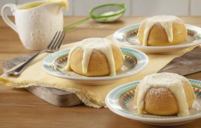 Fotografia em tons de bege com três porções de bolinhos com cobertura em pratinhos coloridos, sobre guardanapo amarelo e tábua de cozinha com um garfo ao lado, pote de cobertura e peneirinha verde ao fundo em bancada de madeira.