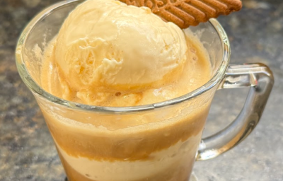 A imagem mostra uma caneca transparente com camadas de uma bebidas em tons de bege, creme e marrom. Uma bola de sorvete e um biscoito Tostines Especiarias Ginger decoram. A caneca está em uma bancada de granito escuro.