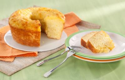 Fotografia em tons de laranja e verde em uma mesa de madeira verde com um pano laranja de bolinhas branca. Ao centro, um prato raso branco com o bolo de macaxeira cortado. Ao lado, um prato branco com borda verde e duas fatias do bolo. Fotografia em tons de laranja e verde em uma mesa de madeira verde com um pano laranja de bolinhas branca. Ao centro, um prato raso branco com o bolo de macaxeira cortado. Ao lado, um prato branco com borda verde e duas fatias do bolo.
