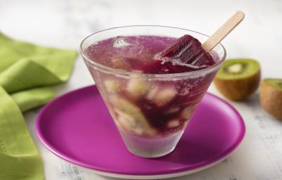 Fotografia em tons de branco, roxo e verde de uma bancada de madeira branca, um paninho verde e um prato redondo roxo, sobre ele um copo redondo de vidro com o drink de picolé. Ao fundo um kiwi cortado ao meio.