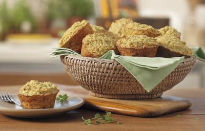 Fotografia em tons de verde em uma bancada de madeira, uma tábua de madeira, uma cesta de vime com vários muffins de aveia e embaixo um paninho verde. Ao lado, um pratinho de sobremesa branco com um muffin em cima dele.