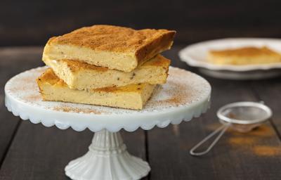 Fotografia em tons de branco, dourado e grafite, com boleira branca contendo três fatias de bolo, uma peneirinha, canela em pó e um prato branco com bolo no entorno, tudo sobre bancada em tons de grafite.