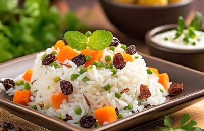 Em um prato de porcelana marrom, há uma porção de arroz branco misturado com cenouras, ervilhas e uvas-passas, decorado com duas folhas de hortelã.