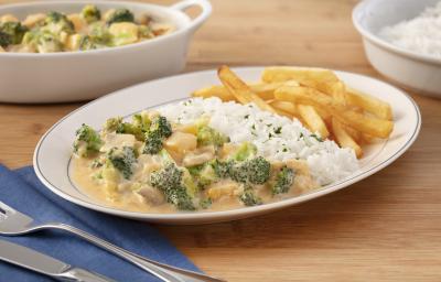 Fotografia em tons de verde em uma bancada de madeira com um guardanapo de pano azul, um prato oval branco no centro com o strogonoff de brócolis, arroz branco e batata frita. Ao fundo, um recipiente com strogonoff de brócolis e outro com arroz. Fotografia em tons de verde em uma bancada de madeira com um guardanapo de pano azul, um prato oval branco no centro com o strogonoff de brócolis, arroz branco e batata frita. Ao fundo, um recipiente com strogonoff de brócolis e outro com arroz.