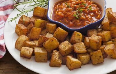 Fotografia em tons de vermelho em uma bancada de madeira escura com um pano branco com listras vermelhas e um prato branco raso grande ao centro com as batatas assadas e um potinho com o molho de tomate picante dentro. Fotografia em tons de vermelho em uma bancada de madeira escura com um pano branco com listras vermelhas e um prato branco raso grande ao centro com as batatas assadas e um potinho com o molho de tomate picante dentro.