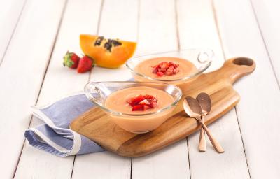 Fotografia em tons de azul e rosa em uma bancada de madeira branca, um paninho azul, uma tábua de madeira com duas taças em cima com creme de papaia dentro delas, decoradas com morangos. Ao fundo mamão e morangos. Fotografia em tons de azul e rosa em uma bancada de madeira branca, um paninho azul, uma tábua de madeira com duas taças em cima com creme de papaia dentro delas, decoradas com morangos. Ao fundo mamão e morangos.