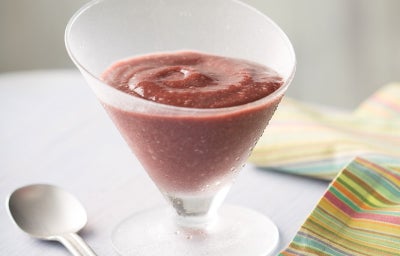Foto de uma taça de vidro fosco com delícia de açaí de cor roxa intensa e textura cremosa, com leve redemoinho na superfície. A taça está sobre mesa clara, ao lado de uma colher prateada e de um guardanapo listrado colorido.