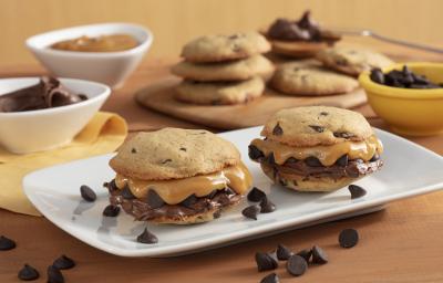 Fotografia em tons de amarelo em uma bancada de madeira com um recipiente branco ao centro com dois cookies recheados com doce de leite e creme de avelã. Ao fundo, potinhos com gotas de chocolate, doce de leite e uma tábua com mais cookies. Fotografia em tons de amarelo em uma bancada de madeira com um recipiente branco ao centro com dois cookies recheados com doce de leite e creme de avelã. Ao fundo, potinhos com gotas de chocolate, doce de leite e uma tábua com mais cookies.