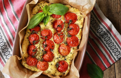 sobre um pano colorido listrado em tons de vermelho e branco está uma forma refratária com uma focaccia assada em tons dourados e toamtes cereja por cima. Folhas de manjericão guarnecem o prato. sobre um pano colorido listrado em tons de vermelho e branco está uma forma refratária com uma focaccia assada em tons dourados e toamtes cereja por cima. Folhas de manjericão guarnecem o prato.