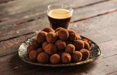 fotografia em tons de marrom e preto de uma bancada marrom vista de frente, contém um prato redondo com trufas com chocolate, e ao fundo um copo com café. fotografia em tons de marrom e preto de uma bancada marrom vista de frente, contém um prato redondo com trufas com chocolate, e ao fundo um copo com café.