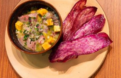 Fotografia vista de cima de um recipiente pequeno com ceviche ao lado de 4 lâminas de batata doce frita, sobre um prato de tom claro apoiado em uma bancada de madeira. Fotografia vista de cima de um recipiente pequeno com ceviche ao lado de 4 lâminas de batata doce frita, sobre um prato de tom claro apoiado em uma bancada de madeira.