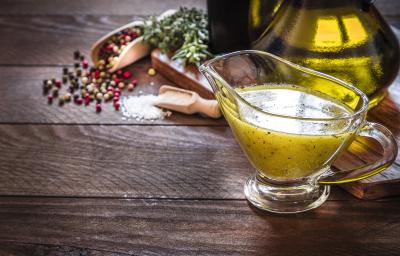 Fotografia de um mini recipiente de vidro com um molho para vinagrete. Ao fundo, pimenta em grãos, sal e azeite, sobre uma mesa de madeira.