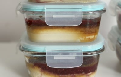 Fotografia de três recipientes de vidro com tampa, um sobre o outro, em uma mesa branca. Dentro dos recipientes tem uma receita de cheesecake.