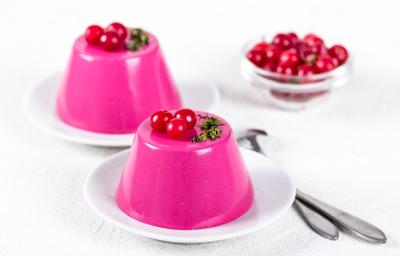 Fotografia de dois pratos pequenos com um manjar na cor rosa em cada, sobre eles, três cerejas para decoração. Ao fundo, várias cerejas em um refratário fundo de vidro pequeno, e ao lado dos pratos, duas colheres de sopa. Tudo está sobre uma mesa branca.