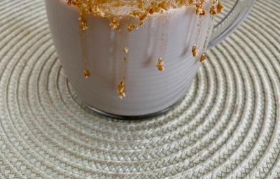 Fotografia em tons de marrom em uma bancada com um pano bege e uma xícara de vidro com a bebida de cappuccino de caramelo salgado dentro. Xícara decorada com calda de caramelo e biscoitos Tostines Especiarias Fotografia em tons de marrom em uma bancada com um pano bege e uma xícara de vidro com a bebida de cappuccino de caramelo salgado dentro. Xícara decorada com calda de caramelo e biscoitos Tostines Especiarias