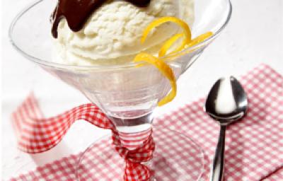 fotografia em tons de branco, vermelho e marrom de uma bancada branca vista de frente, contém um pano xadrez em tons de vermelho e branco. Uma taça transparente com uma bola de sorvete de creme com calda de chocolate