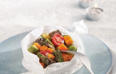 Fotografia em tons de azul em uma mesa com uma toalha branca de renda, um prato branco e azul claro em cima dela e o papillote de legumes (abobrinha, cenoura, tomate) em cima do prato azul, enrolado em um papel manteiga.