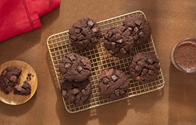 Fotografia de uma grelha quadriculada ao lado de um copo de vidro com leite e nescau e um mini apoio circular de madeira com um cookie ao meio. Sobre a grelha tem sete cookies de nescau extra cacau com gotas de chocolate.